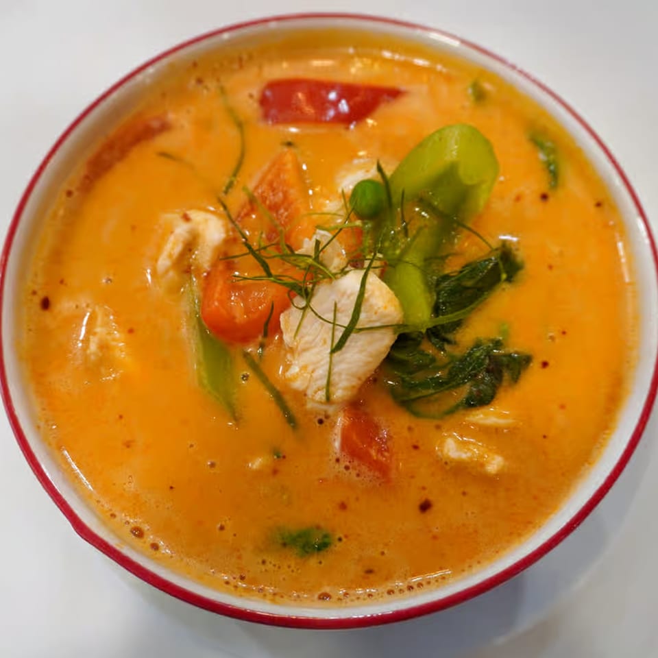 Close-up of a vibrant orange Thai curry with chicken, tomatoes, and green herbs in a white bowl.
