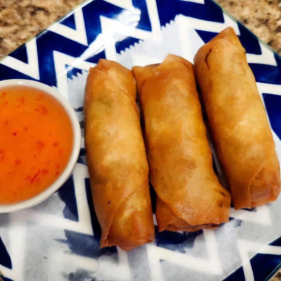 Three crispy golden-brown Thai spring rolls with a side of sweet chili dipping sauce on a blue and white plate.
