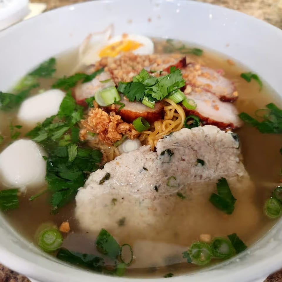 Close-up of Thai noodle soup with pork, fish balls, egg, and fresh herbs in a white bowl.