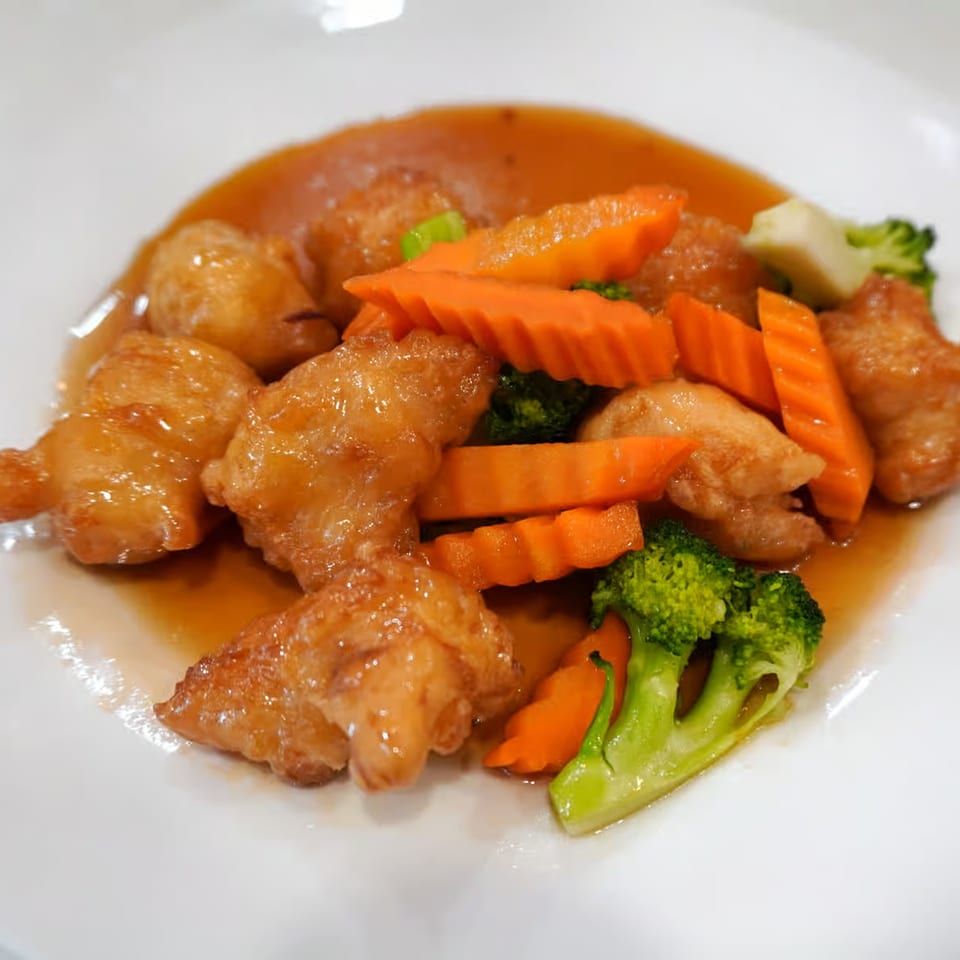 Sweet and sour chicken with broccoli and carrots in a rich orange sauce on a white plate.