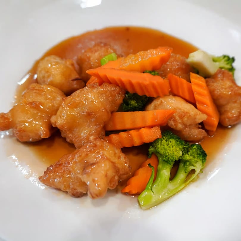 Sweet and sour chicken with broccoli and carrots in a rich orange sauce on a white plate.